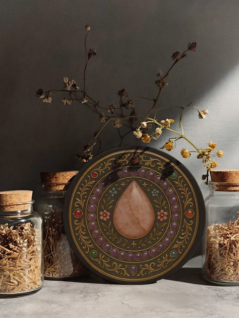 Round ceramic drink coaster featuring a detailed rose quartz-inspired teardrop surrounded by floral and gemstone baroque patterns. Black background with jewel-toned accents and gold scrollwork. The coaster has a non-slip cork base and measures approximately 4 inches in diameter. Perfect for hot or cold beverages, this witchy decor piece enhances any coffee cart, altar, or side table. Suitable for use in whimsigoth, gothic, or celestial-themed spaces.